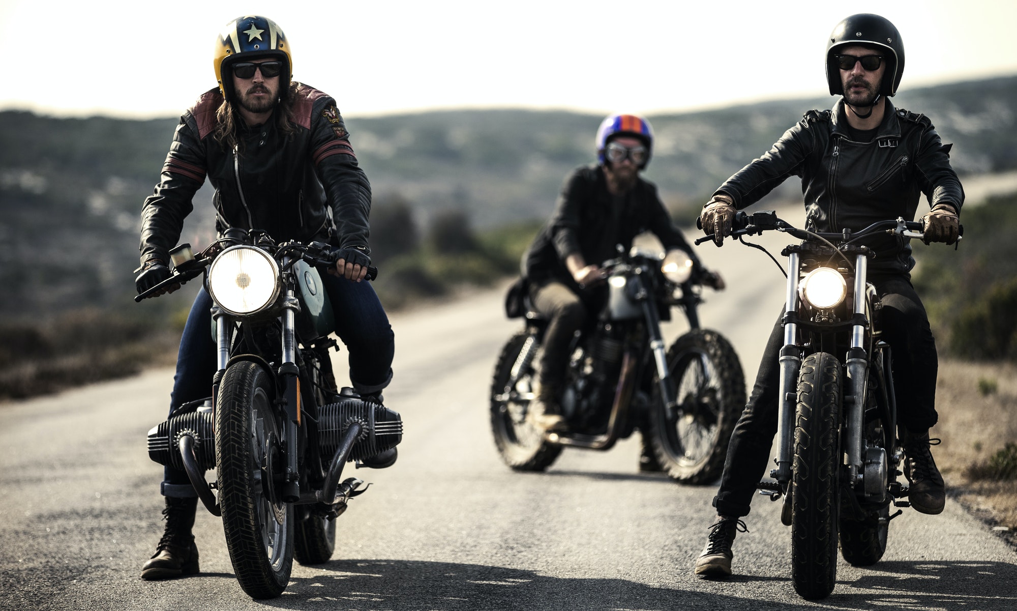 three-men-wearing-open-face-crash-helmets-and-goggles-sitting-on-cafe-racer-motorcycles-on-a-rural.jpg